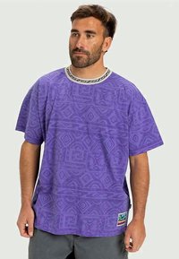 Camisa de manga corta morada con patrones geométricos texturizados, cuello redondo con acentos en blanco y negro, y un parche con logo en el dobladillo.