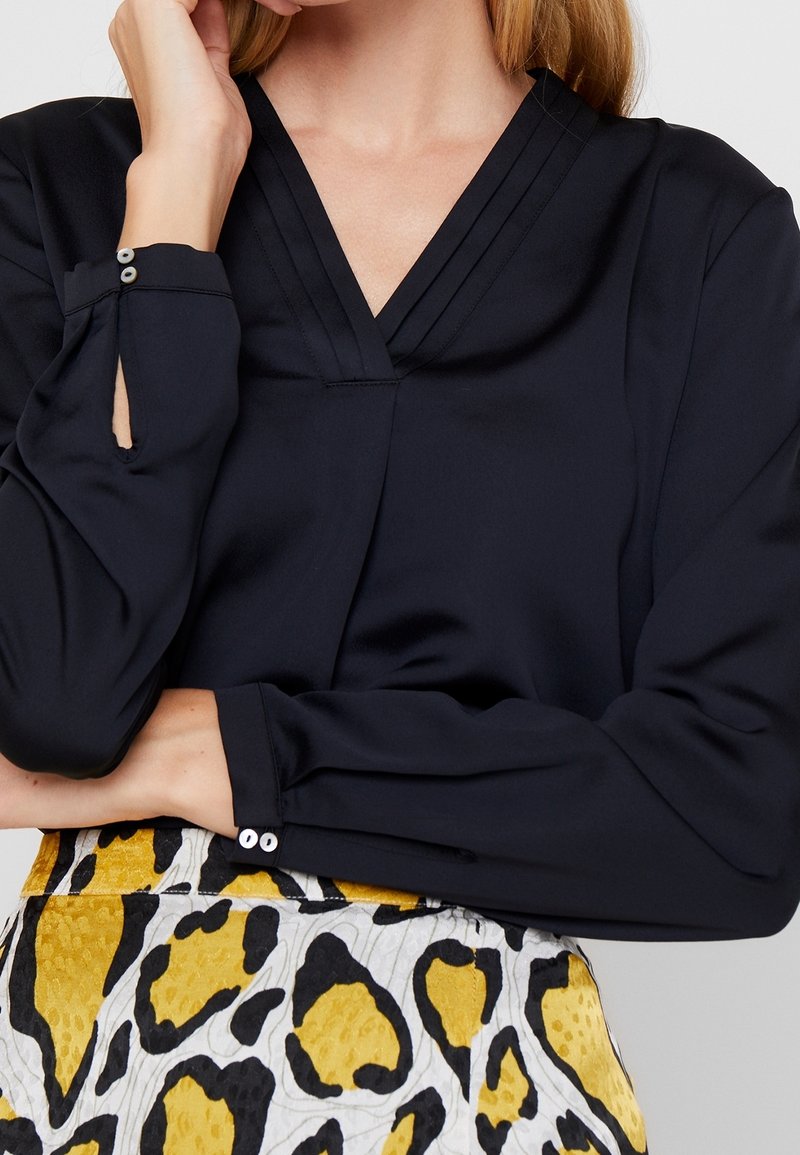 Marineblauwe satijnen blouse met een V-hals en lange mouwen, voorzien van een subtiel geplooid detail en knoopmanchetten. Gecombineerd met een rok met een geel en zwart patroon.