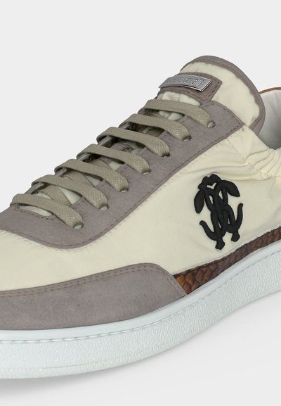 Αθλητικό παπούτσι σε εκρού και γκρι χρώμα με γκρι κορδόνια, μαύρο κεντημένο λογότυπο στο πλάι και καφέ υφασμάτινη λεπτομέρεια πάνω από τη λευκή σόλα.