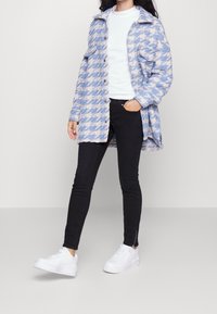 Persona vestida con un abrigo de houndstooth en azul claro y blanco, camisa blanca, pantalones negros y zapatillas blancas, de pie con una mano en el bolsillo.