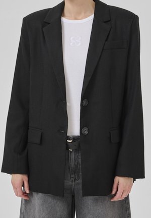 Persona que lleva una blazer negra sobre una camisa blanca acanalada y vaqueros grises desteñidos con un cinturón negro, de pie frente a un fondo liso.