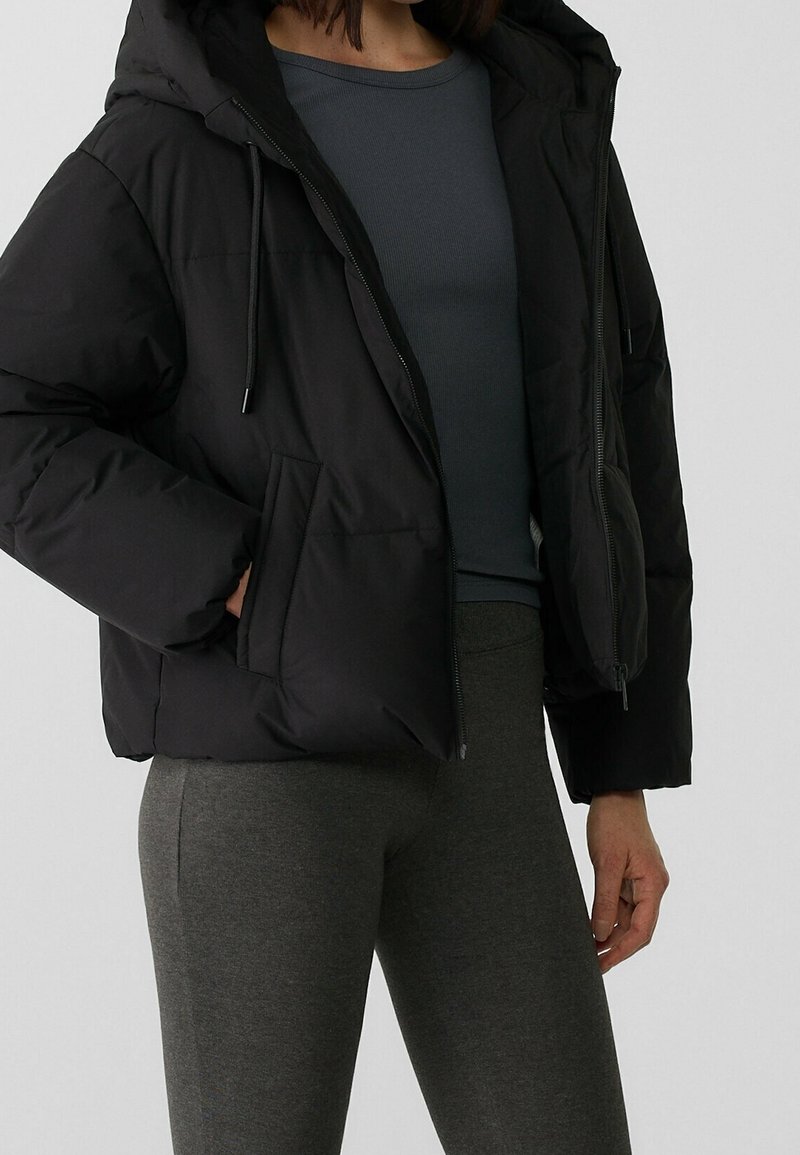 Femme portant une doudoune noire à capuche, un t-shirt à manches longues gris foncé et un pantalon gris ajusté, posant devant un fond uni.