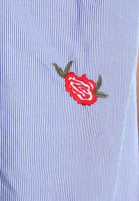 Niebiesko-biała tkanina w paski z haftowanym czerwonym różą i zielonymi liśćmi. Tkanina jest gładka o wyraźnej teksturze.