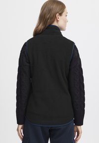 Gilet en polaire noire avec des manches en tricot câble bleu marine, un col montant et des bordures bleu le long des épaules, avec une finition texturée sur les manches.