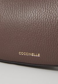Brown textured leather handbag with gold "COCCINELLE" logo. Features a smooth black seam at the base, highlighting the elegant design.