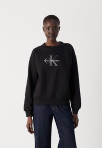 Čierna mikina s okrúhlym výstrihom, dlhými rukávmi a rebrovanými manžetami. Obsahuje strieborné logo Calvin Klein a potlačené "ck" uprostred.