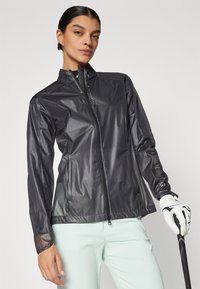 Vattentät svart zip-up jacka med slim fit, med hög krage och genomskinliga ärmar, i kombination med mintfärgade byxor och en golfklubba.