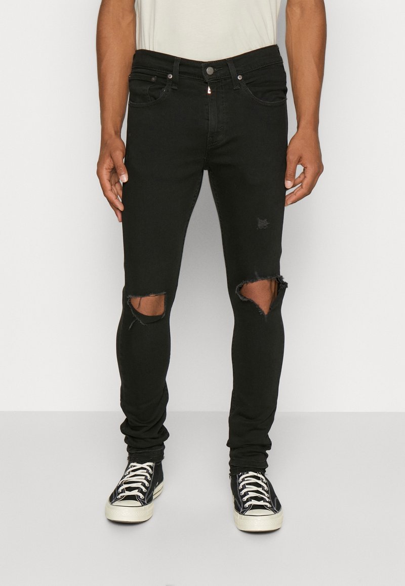 Homme portant un jean skinny noir déchiré et des baskets noires et blanches debout devant un fond uni.
