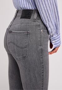 Jeans de mezclilla gris con una cintura alta, bolsillos delanteros y traseros, y un parche negro de la marca "Lee" en la cintura. Detalle de desgaste sutil.