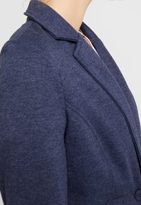 Blazer azul marino con tejido de punto texturizado, solapas con muesca y cierre de un solo botón. Ajuste entallado con una sutil forma en los hombros.