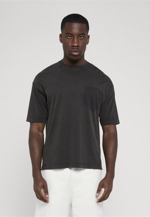 Man met een effen zwarte korte mouw T-shirt met borstzakje en witte broek, staand tegen een effen lichte achtergrond.