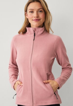 Vrouw draagt een roze fleecejack met ritssluiting, met handen in de zakken, staand voor een effen lichte achtergrond.