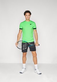 Grønn idretts t-skjorte med svarte striper, parret med mørke shorts. Holder en tennisracket, iført hvite joggesko og sokker.