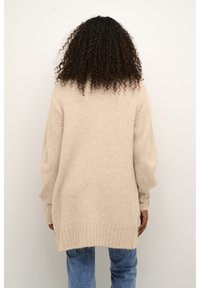 Beige, oversized kötött pulóver bordás passzéval és szegéllyel, laza szabással és puha textúrával. Kék farmernadrág fölött viselve, hátulról látható.