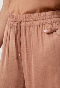 Perzikkleurige, lichte broek met een elastische tailleband en een trekkoord. Bevat twee zijzakken en een soepele, textuurstof.