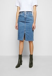 Woman wearing mid-length blue denim skirt with front slit, white tucked-in t-shirt, and dark green ankle boots, standing against a plain background.