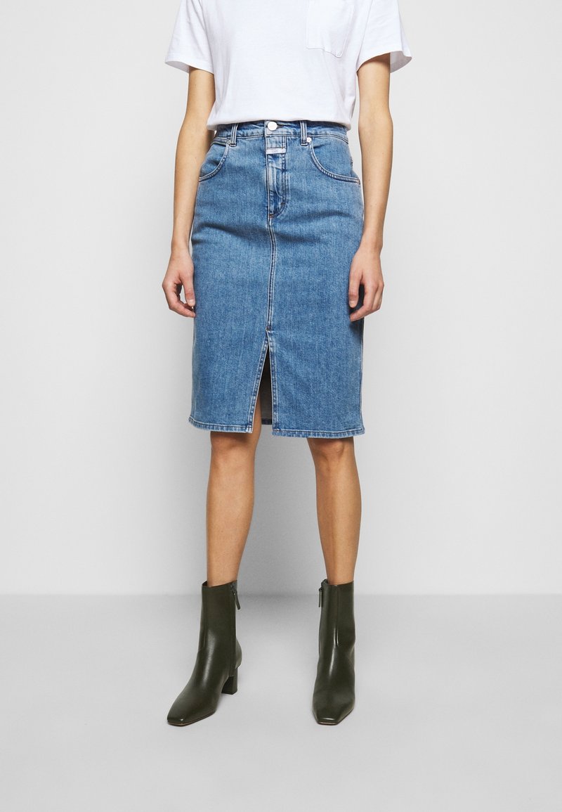 Woman wearing mid-length blue denim skirt with front slit, white tucked-in t-shirt, and dark green ankle boots, standing against a plain background.