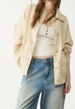 Vrouw draagt een beige jasje met knopen over een cropped wit geribd topje met knopen, gecombineerd met een high-waisted blauwe spijkerbroek en ringen aan haar vingers.