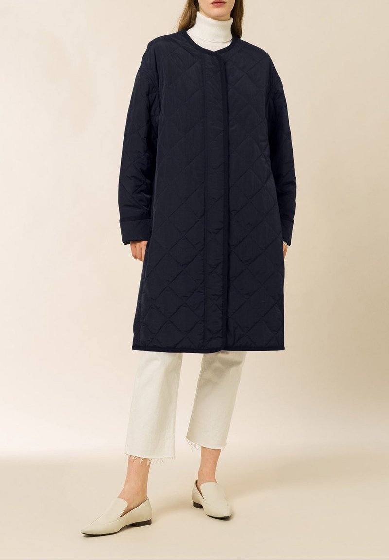 Abrigo largo acolchado azul marino con cuello redondo, cierre frontal, bolsillos laterales y corte relajado, combinado con jeans blancos recortados y zapatos beige.
