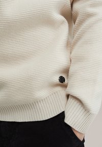 Pulover tricotat de culoare crem, cu manșete și tiv în dentare. Prezintă un mic buton cu logo negru pe lateral pentru detaliu.