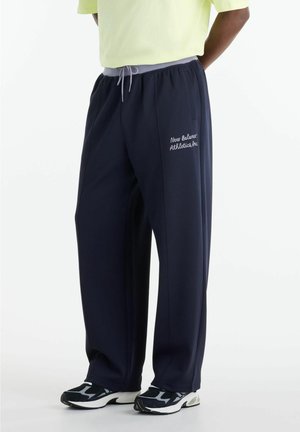 Person står iført marineblå New Balance træningsbukser med grå snor og sorte og hvide sportssko.
