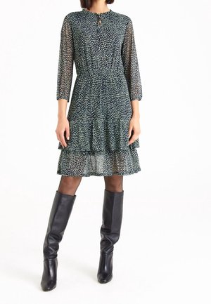 Woman wearing a patterned knee-length dress with three-quarter sheer sleeves and black knee-high leather boots, standing on a white background.