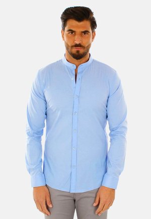 Uomo con capelli scuri e barba che indossa una camicia azzurro chiaro con colletto mandarino abbottonata e pantaloni grigi, in piedi davanti a uno sfondo uniforme.