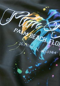 Čierna textília so živými škvrnami modrej, žltej a ružovej farby. Text znie "Palm Beach, Florida" a geografické súradnice.