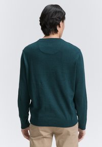 Suéter verde oscuro con cuello redondo y mangas largas. Textura suave, dobladillo y puños acanalados, y un sutil diseño circular en la parte posterior.