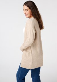 Beige vest met lange mouwen, open voorkant en twee zijzakken. De gebreide textuur en geribbelde zoom creëren een zachte, gezellige uitstraling.