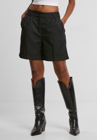 Černé na míru šité šortky s záhyby, s bočními kapsami a zapínáním na knoflík. Kombinováno s elegantními černými vysokými botami.