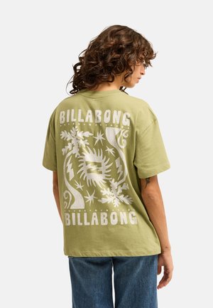 Personne aux cheveux bouclés portant un t-shirt Billabong vert clair avec un motif graphique blanc et un texte dans le dos, associé à un jean bleu.