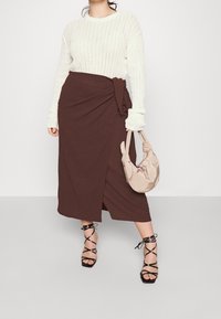 Bruine wikkelrok met strikdetail, gecombineerd met een getextureerde witte trui. Zwarte hakken met bandjes en een beige tas maken de outfit compleet.