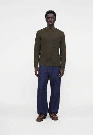 Uomo in piedi rivolto frontalmente, indossa un maglione a coste verde oliva, jeans blu larghi e scarpe di pelle marroni su sfondo bianco semplice.