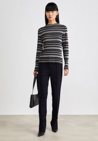 Schwarz-weiß gestreiftes Langarmshirt mit rundem Ausschnitt, kombiniert mit enganliegenden schwarzen Hosen und einer schwarzen Tasche, vor einem neutralen Hintergrund.