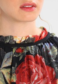 Blomklänning med stora röda rosor och gröna blad på en svart bakgrund. Volangdesign vid halsringningen med svart kant. Mjuk, lättviktig tyg.