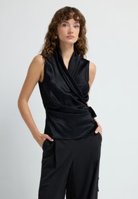 Top sin mangas de satén negro con escote drapeado y diseño envolvente, que presenta un detalle anudado en la cintura y un acabado liso y brillante.