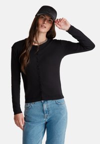 Zwarte geribde cardigan met een knoopsluiting aan de voorkant, lange mouwen en een aansluitende taille. Gecombineerd met lichtblauwe jeans en een zwarte pet.