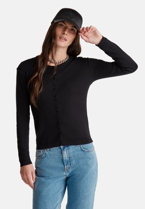 Zwarte geribde cardigan met een knoopsluiting aan de voorkant, lange mouwen en een aansluitende taille. Gecombineerd met lichtblauwe jeans en een zwarte pet.