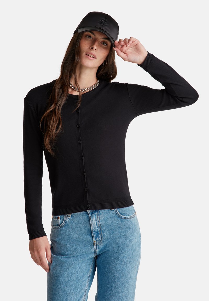 Zwarte geribde cardigan met een knoopsluiting aan de voorkant, lange mouwen en een aansluitende taille. Gecombineerd met lichtblauwe jeans en een zwarte pet.