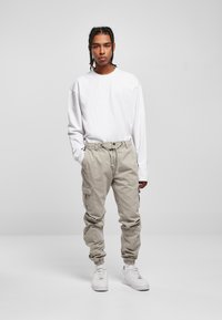 Leichte, langärmlige weiße Bluse kombiniert mit beigefarbenen Cargo-Hosen mit elastischen Bündchen und Seitentaschen, ergänzt durch weiße Sneakers.