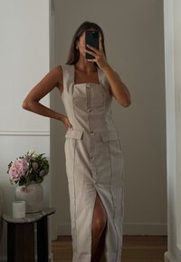 Beige linnedragt med firkantet halsudskæring, knapper ned langs fronten, sideslids og to forlommer. Glat tekstur og skræddersyet pasform.