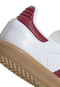 Baskets blanches avec des accents rouges ; tige en cuir lisse, semelle en caoutchouc texturé avec un motif en chevrons, talon renforcé avec des rainures.