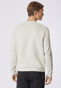 Felpa color beige chiaro con scollo rotondo e maniche raglan. Presenta orlo e polsini a costine. Jeans in denim nero visibili al bordo inferiore.