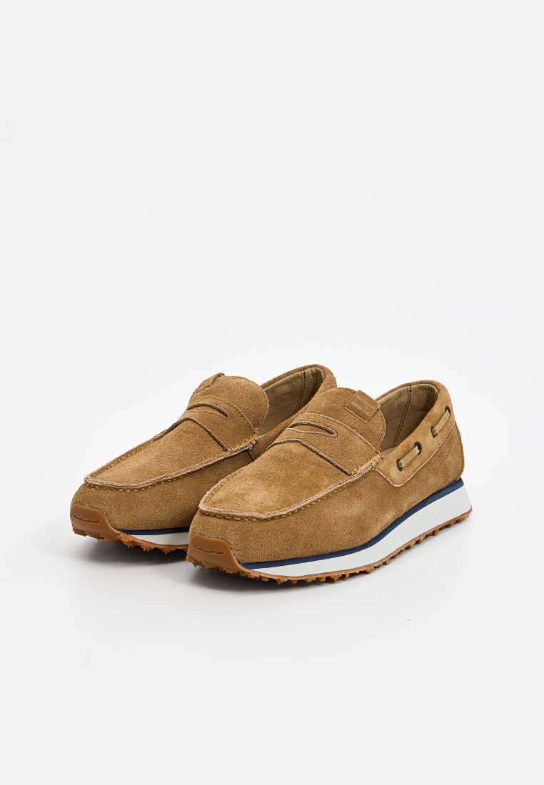 Mocassins en daim marron clair avec un bout arrondi, semelle bicolore blanche et marine, couture décorative et accents latéraux subtils.