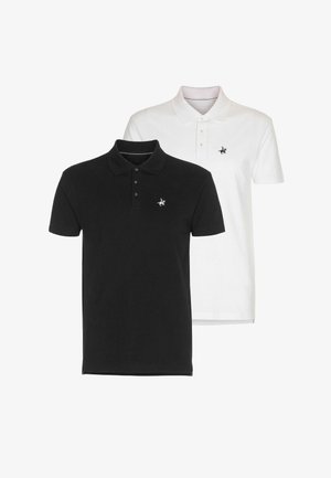 Homme debout portant un polo blanc, un short bleu foncé, des chaussettes noires et des chaussures noires avec des tatouages visibles sur son bras droit.