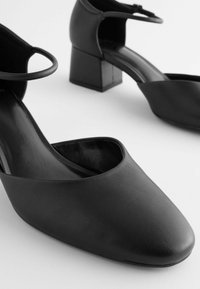 Czarne skórzane buty z odkrytym piętą, kwadratowymi obcasami, gładką fakturą i zakrzywionym wzorem na cholewce, tworzące minimalistyczny wygląd.