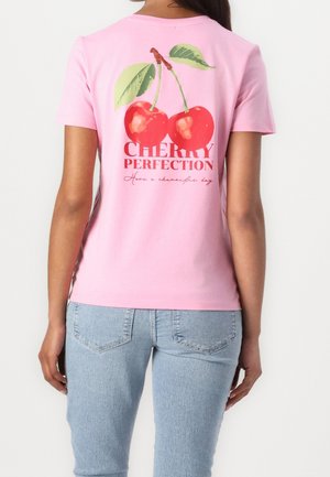 Frau trägt ein rosa T-Shirt mit zwei roten Kirschen und dem Text "CHERRY PERFECTION Have a cherryful day" auf dem Rücken, kombiniert mit hellblauen Jeans.