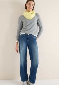 Pull gris à manches longues, foulard jaune à motifs autour du cou, jean bleu à jambes larges, et baskets argentées sur un fond clair.