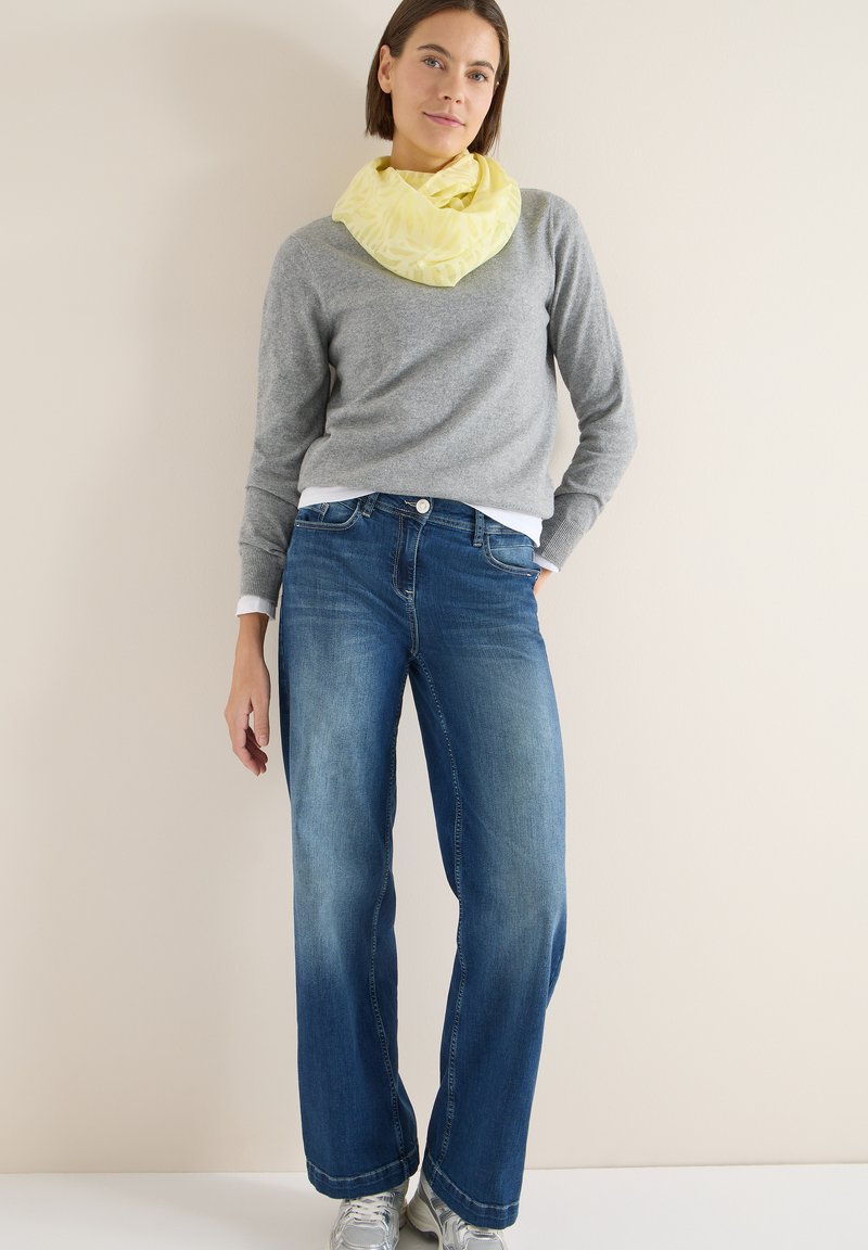 Pull gris à manches longues, foulard jaune à motifs autour du cou, jean bleu à jambes larges, et baskets argentées sur un fond clair.